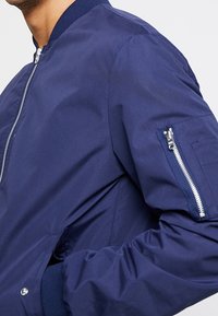 Námořnická bomber bunda se zipem vpředu, žebrovaným límcem a manžetami, s kapsou na zip na rukávu a hladkou, lehkou tkaninou.