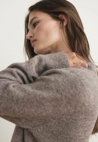 Brauner Strickpullover mit gerippter Textur und überschnittenen Schultern. Das Model hält sanft ihre Schulter, um den weichen Stoff zu präsentieren.