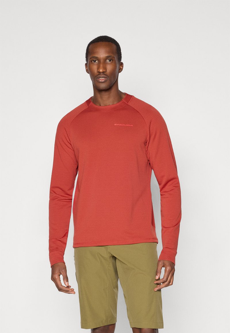 Rød langermet skjorte med ribbet tekstur, rund hals og subtil merking. Brukt med lysegrønne shorts. Enkelt design og tettsittende fasong.