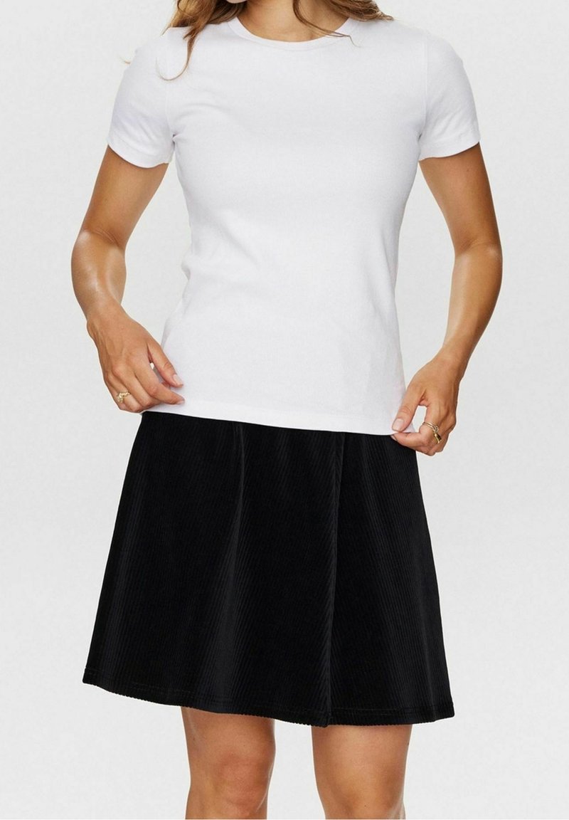 Femme portant un t-shirt blanc uni à manches courtes et une jupe noire longueur genou, tenant l'ourlet du t-shirt avec les deux mains.
