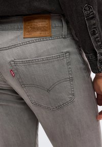 Κοντινό πλάνο πίσω τσέπης γκρι τζιν Levi's με καφέ δερμάτινο ταμπελάκι επωνυμίας και κόκκινη ετικέτα λογότ υπου, φορεμένο με σκούρο τζιν μπουφάν.