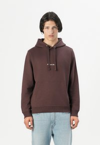 Brown cotton hoodie with a front pocket, adjustable drawstrings, and a small logo on the chest, paired with light blue jeans.