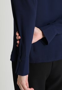 Blouse à manches longues bleu marine avec une texture lisse, dotée de poignets boutonnés et d'un ourlet droit, partiellement rentrée derrière une main.