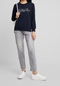 Felpa blu navy con design in scrittura bianca, abbinata a jeans denim grigi chiari e sneakers bianche con piattaforma. Estetica semplice e casual.