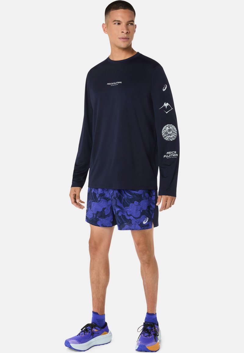 Chemise de sport à manches longues bleu marine avec logos et motifs blancs, associée à un short violet à motifs. Chaussures de sport avec des accents bleus et orange.