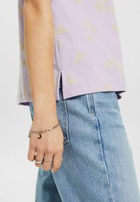 Světle fialový krátký rukáv top s žlutým květinovým vzorem, s bočními rozparky. V kombinaci s modrými džínami a stříbrnými šperky na ruce.