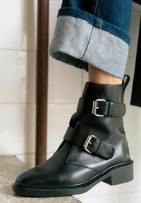 Stivali alla caviglia in pelle nera con due cinturini a fibbia e punta tonda, abbinati a jeans in denim blu con orlo arrotolato. Texture liscia e suola robusta.