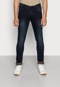 Mørkeblå denim jeans med slim fit, med fem lommer, glidelås og oppbrettede ermer. Kombinert med hvite joggesko.