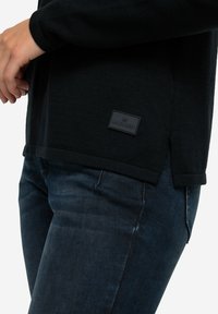Maglione nero a maglia con spacchi laterali e un'etichetta strutturata; abbinato a jeans in denim scuro, che evidenziano la forma e la vestibilità del capo.