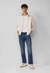 Hellbeige Zip-Jacke aus Fleece über einem weißen T-Shirt, kombiniert mit blauen Jeans und cremefarbenen Sneakern mit braunen Akzenten.