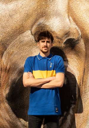 Blauwe en gele polo met kraag en korte mouwen, voorzien van een klein logo. Model staat tegen een gestructureerde bruine muur.