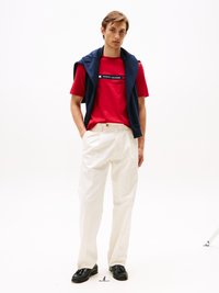 Rotes T-Shirt mit weißem "TOMMY HILFIGER"-Grafik, kombiniert mit weißen Hosen. Ein marineblauer Pullover liegt über den Schultern. Schwarze Slipper werden getragen.
