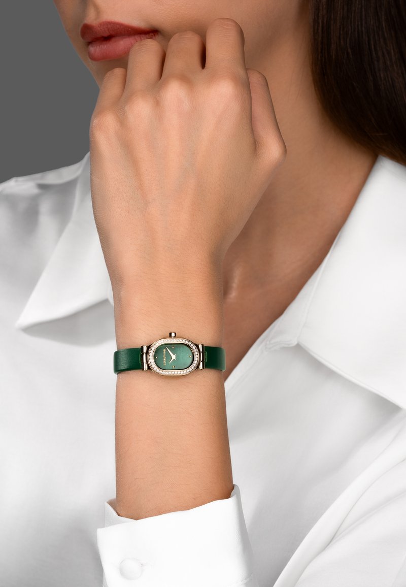 Mujer con un reloj de pulsera de correa de cuero verde, con esfera ovalada de esmeralda y bisel engastado en diamantes, en su muñeca izquierda, mano cerca del rostro.