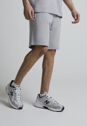 Mann iført lysegrå shorts og hvite New Balance-sneakers, stående mot en ensfarget lys grå bakgrunn.