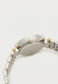 Stříbrné a zlaté tónové náramkové hodinky s hladkým kulatým pouzdrem, třemi knoflíky a spojeným kovovým páskem s kartáčovaným a leštěným povrchem.