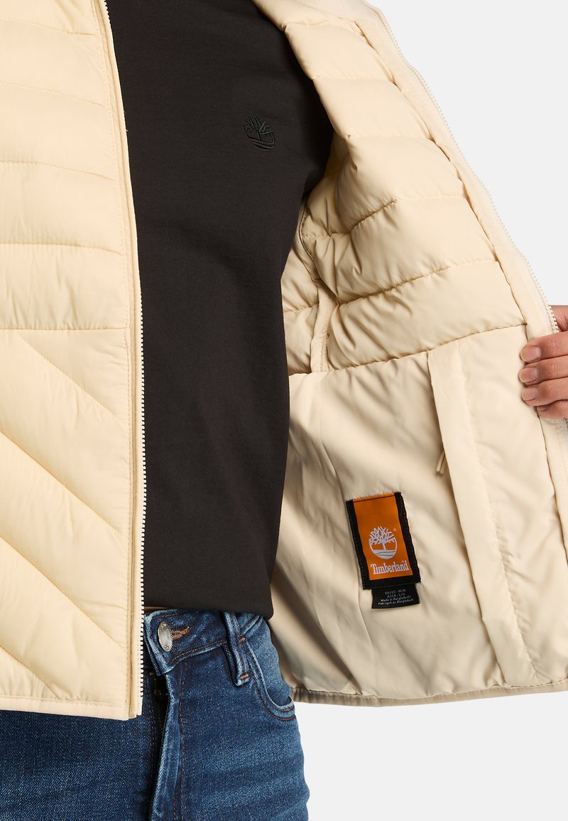 Giacca piumino beige con chiusura a zip, texture trapuntata e tasca interna con logo Timberland su un'etichetta arancione.