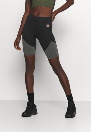 Atletisk kvinde iført sorte og grå cykelshorts, grøn cropped tanktop, sorte sokker og sorte træningssko, stående mod en hvid baggrund.