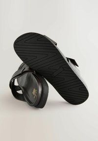 Sandale noire avec une tige en cuir texturé et une semelle en EVA, présentant un design minimaliste et un détail de profil prononcé sur la semelle extérieure.