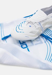 Bijele nogometne kopačke s plavim akcentima, s teksturiranim uzorcima i trakicom za povlačenje, odložene na bijelu torbu s plavim crtama.