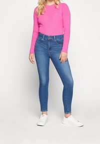 Jeans de tiro alto en azul con un diseño ajustado, combinados con un suéter de canalé en rosa brillante. Zapatillas blancas completan el conjunto.