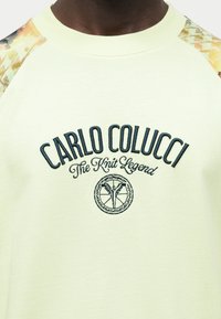 Κοντινή λήψη ανοιχτόκίτρινου φούτερ με το κείμενο «Carlo Colucci The Knit Legend» και κυκλικό λογότυπο στο στήθος, με σχέδιο στα μανίκια σε κίτρινη και μπεζ απόχρωση.
