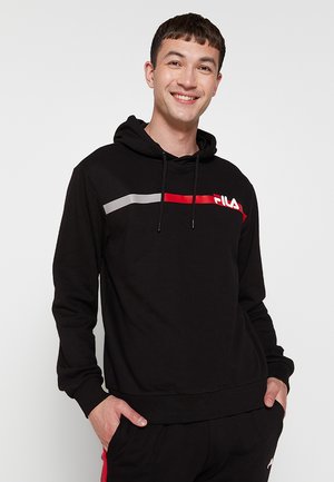 Junger Mann lächelt, trägt einen schwarzen FILA Hoodie mit roten und weißen Streifen und schwarze Hosen, Hände in den Taschen, vor einem einfarbigen hellen Hintergrund.