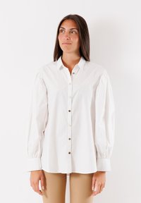 Camicia in cotone bianco con bottoni sul davanti, colletto a punta e maniche a sbuffo. Presenta bottoni beige e polsini aderenti. Texture liscia.