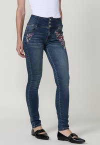 Jeans de mezclilla azul de tiro alto con bordado floral en el bolsillo derecho, cierre de cinco botones y diseño de pierna ajustada.