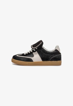 Flacher Sneaker mit schwarzen und weißen strukturierten Einsätzen, weißen Schnürsenkeln, umgelegter Zunge und brauner Gummisohle, im Profil vor weißem Hintergrund gezeigt.