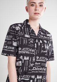 Maglietta nera a maniche corte con diverse grafiche e testi bianchi, realizzata in nylon, con una vestibilità rilassata e dettaglio della tasca sul petto.