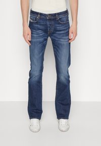 Jeans de mezclilla, de un azul oscuro con un lavado desvanecido. Cuenta con cinco bolsillos, costuras plateadas y un cierre de botón. Diseño de pierna recta. Se llevan zapatillas blancas.