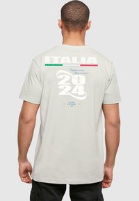 T-shirt in cotone grigio chiaro con il testo "ITALIA", accenti verde e rosso, e "2024" in bianco in grassetto. Dettagli stampati includono stelle e frasi.
