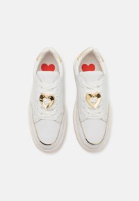 White sneakers with gold accents, heart-shaped decoration on laces, rubber sole, and red heart pattern on insoles. Smooth texture.