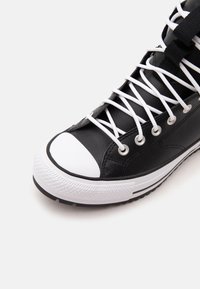 Μαύρο αθλητικό παπούτσι ψηλού τύπου με λευκή λαστιχένια μύτη, λευκά κορδόνια και λευκή λαστιχένια σόλα σε ανοιχτό φόντο.