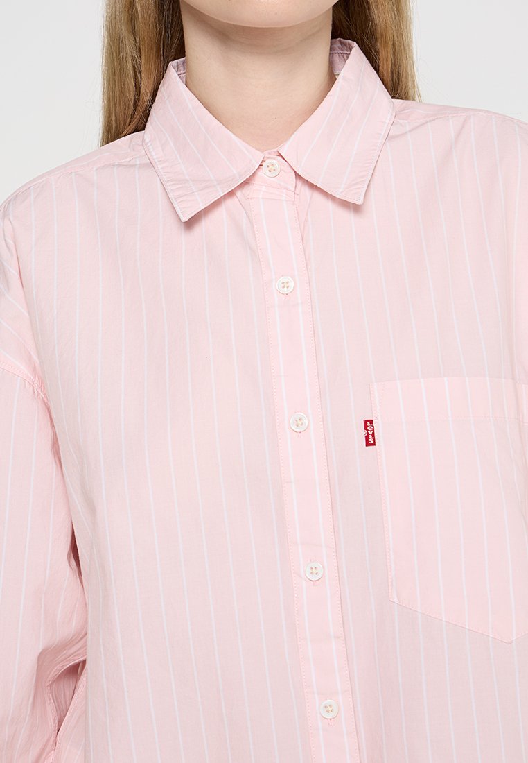 Chemise boutonnée rose pâle avec de fines rayures blanches, col pointu, poche frontale et une petite étiquette logo rouge sur le côté gauche.