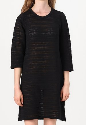 Vrouw draagt een zwarte, halfdoorzichtige jurk met lange mouwen en horizontale kanten strepen, staand tegen een effen witte achtergrond.