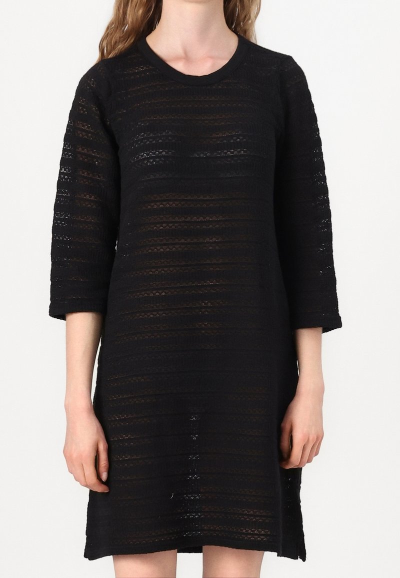 Femme portant une robe noire semi-transparente à manches longues avec des rayures horizontales en dentelle, debout devant un fond blanc uni.