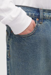 Χέρι μέσα στην μπροστινή τσέπη μπλε τζιν παντελονιού που φοριέται με λευκό μακρυμάνικο πουκάμισο ορατό στον καρπό.