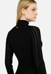 Pull noir côtelé avec un col haut, présentant un design de tricot texturé et une subtile tresse sur les côtés. Bouton doré en accent au dos.