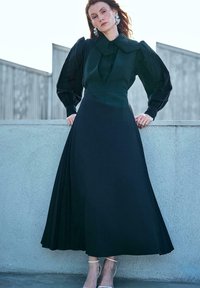 Femme en longue robe noire à manches bouffantes et grand col, debout contre un mur en béton, portant des talons blancs et de grandes boucles d’oreilles.