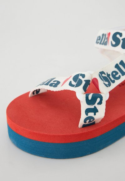 Sandalia de plataforma roja y azul con tiras blancas que presentan el texto "Stella". Textura suave, diseño de soporte y capas de color en contraste.