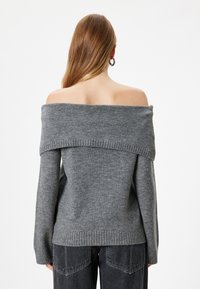 Grau off-shoulder Pullover aus weichem Strick, mit einem gerippten Halsausschnitt und Bündchen. Darunter sind ausgefranste schwarze Jeans sichtbar.