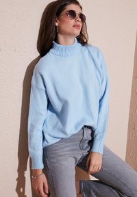 Gaišzils, pārlieku liels turtlenecks džemperis no maigas dzijas, kombinēts ar gaiši pelēkām džinsu biksēm. Aksesuāri: apaļas saulesbrilles un sudraba aproce.