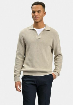 Beige strikket pullover med polo krave og ribbet tekstur, båret over en hvid t-shirt. Modellen har mørke bukser på, med hænderne i lommerne.