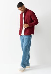 Chemise bordeaux boutonnée avec poche, associée à un jean bleu clair et des baskets blanches. Coupe décontractée, confortable, et tissu texturé.