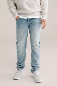 Světle modré džíny z denimu s volnějším střihem, nošené s béžovým prošívaným svetr a bílými teniskami se světle zelenými akcenty.