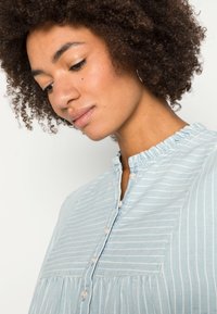 Blusa risca azul clara com decote plissado, fecho de botão e padrão franzido. Feita de um tecido suave com textura subtil.