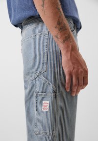 Torso y mano de un hombre visibles, vistiendo pantalones a rayas azules con un bolsillo trasero y un antebrazo tatuado contra un fondo sencillo.