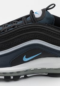 Kengät Nike Air Max 97, joissa on kerroksittain mustaa ja tummansinistä kangasta, vaaleanharmaita yksityiskohtia, sininen brodeerauspistete logo ja läpinäkyvä ilmatyyny.