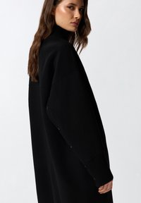 Maglione a collo alto nero, oversize, con maniche lunghe, dettagli con bottoni lungo il lato e polsini a costine, realizzato in un morbido materiale testurizzato.
